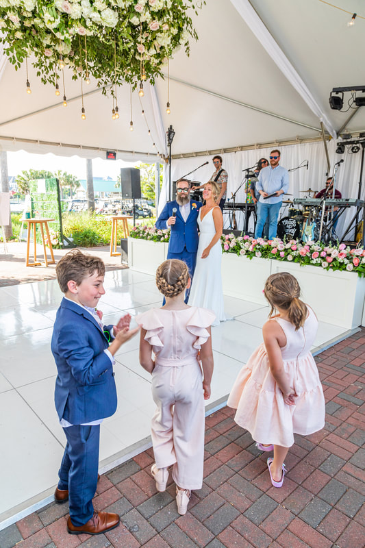 Knot Too Shabby Events, NC Wedding Planner, NC Wedding Coordinator, Bald Head Island Wedding, Summer Wedding, Pink, Blue, White, Coastal Chic, Elegant, Coral, Teal, Bride and Groom on dance floor staring happily at kids. Kids are two flower girls under the age of 10 wearing light pink dresses, and 1 ring bearer wearing a matching blue tux as the groom. Bride and groom are standing on white dance floor under a tent with a stage lined with colorful pink flowers in the background. 