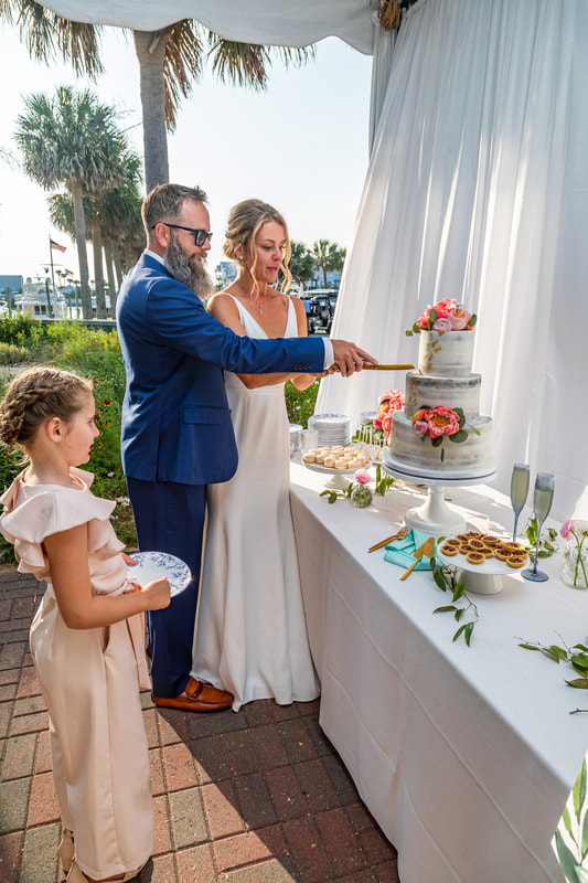 Knot Too Shabby Events, NC Wedding Planner, NC Wedding Coordinator, Bald Head Island Wedding, Summer Wedding, Pink, Blue, White, Coastal Chic, Elegant, Coral, Teal, Bride wearing a long white dress, and groom wearing a blue tux, are holding a cake knife together cutting a 3-tiered white cake decorated with pink flowers. Cake is on a white round cake stand on a 6 foot rectangular dessert table with a white linen covering it. Also on the dessert table are a multitude of desserts.