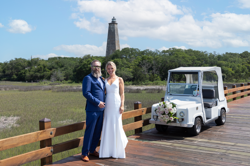 Knot Too Shabby Events, NC Wedding Planner, NC Wedding Coordinator, Bald Head Island Wedding, Summer Wedding, Pink, Blue, White, Coastal Chic, Elegant, Coral, Teal, Bride and Groom standing together on a small wooden bridge. Groom is in a deep blue tux while the bride is in a long white dress. Beside them is a white rolls Royce golf cart decorated with flowers and in the background is the bald head lighthouse. 
