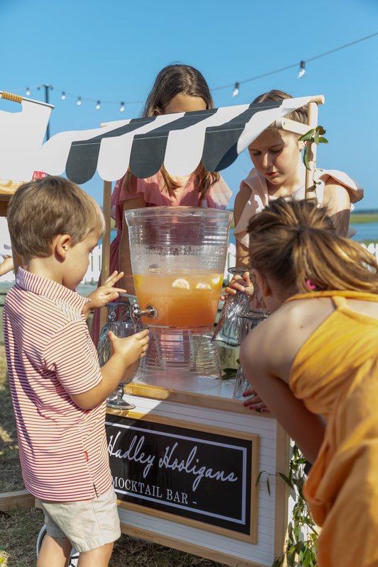 Knot Too Shabby Events, NC Wedding Planner, NC Wedding Coordinator, Bald Head Island Wedding, Summer Wedding, Pink, Blue, White, Coastal Chic, Elegant, Coral, Teal, A child size drink cart holding a clear pitcher full of a non-alcoholic beverage with kids around filling their ice cream cone clear shaped glasses. 