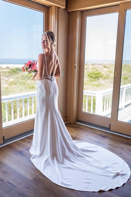 Knot Too Shabby Events, NC Wedding Planner, NC Wedding Coordinator, Bald Head Island Wedding, Summer Wedding, Pink, Blue, White, Coastal Chic, Elegant, Coral, Teal, Bride in an elegant white dress with a circle train, peering out over the sand dunes from a set of corner floor-length windows of a beach house, holding her bouquet of colorful flowers. 