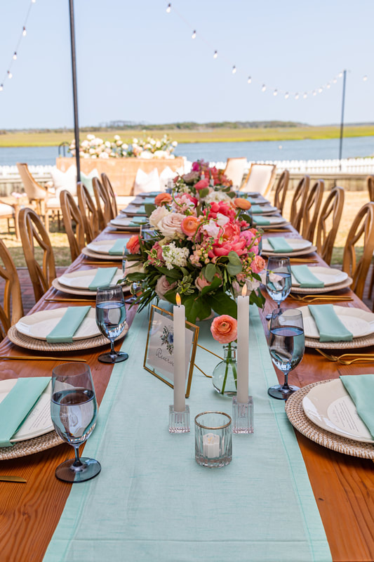 Knot Too Shabby Events, NC Wedding Planner, NC Wedding Coordinator, Bald Head Island Wedding, Summer Wedding, Pink, Blue, White, Coastal Chic, Elegant, Coral, Teal, A close up of an light wooden farm table with ice blue table runner ice blue napkin, white round plates, white led taper candles, clear bud vases, and bouquet of colorful pink flowers. 