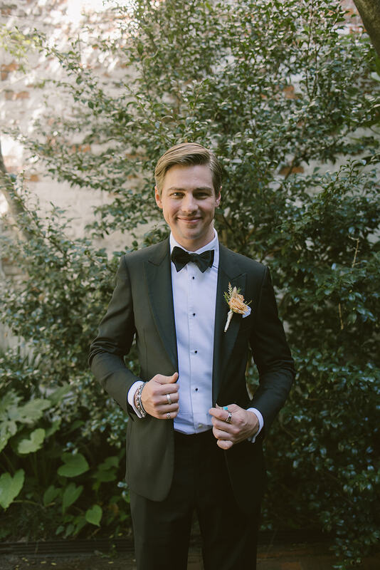 An over joyful groom in a classic black suit and crisp white shirt, featuring a sophisticated dusty pink rose boutonniere, getting ready for his wedding day.