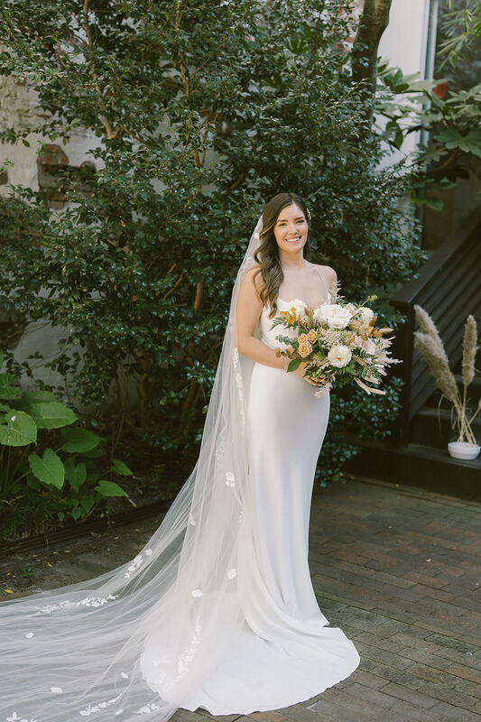A radiant bride graced in a detailed veil, holding a bouquet of coastal-inspired flowers, featuring warm hues and delicate blooms that complement the seaside ambiance.