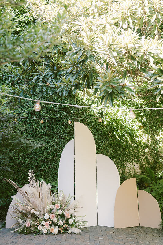A close-up of the ceremony arbor which consists of different heights of beige and pink boards aligned together to form a sort of screen. A large floor plaque of an assortment of boho, earthy flowers on the bottom left of the arbor.   