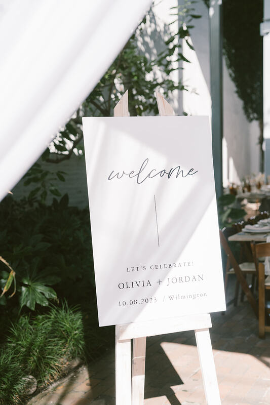 A white cardboard welcome sign with black script writing and on a natural wood easel, at the front of the wedding venue, welcoming all guests to the wedding of Olivia and Jordan. 