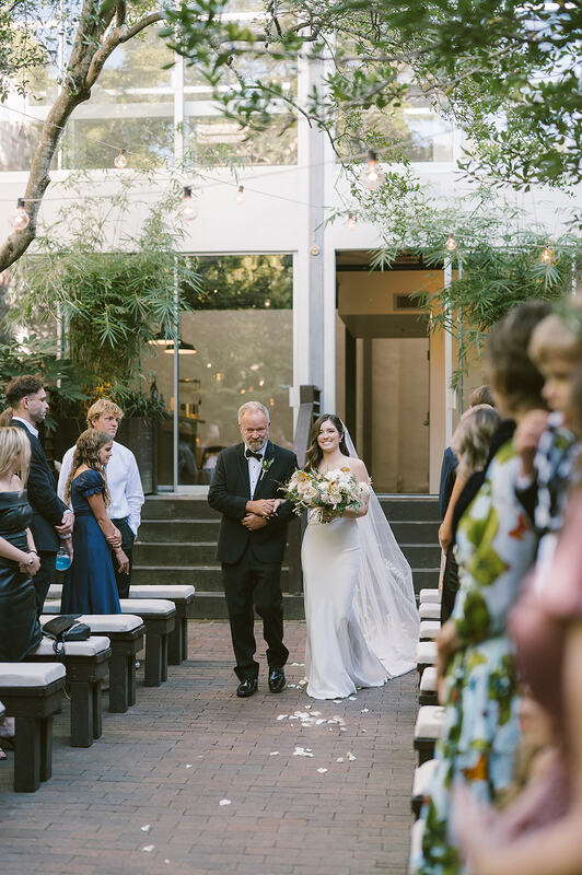 A breathtaking moment as the bride, adorned in a stunning, sleek wedding gown, holding a bouquet of coastal flowers, walks down the aisle escorted by her father. The love and anticipation are palpable, creating a timeless and heartwarming scene looked on by standing guests.