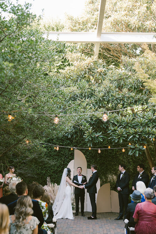 A magical outdoor wedding ceremony set in a rustic, earthy venue. The bride, wearing a beautiful gown, holds hands with the groom amidst a backdrop of nature. Bridesmaids don terracotta dresses, while groomsmen don elegant black tuxedos, creating a harmonious blend of earthy tones and classic sophistication.
