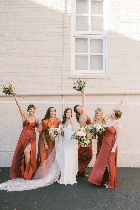 A joyful bridal party on the wedding day, with the bride and bridesmaids wearing chic terracotta-colored dresses, each holding vibrant bouquets in coordinated hues, creating a harmonious and stylish ensemble.