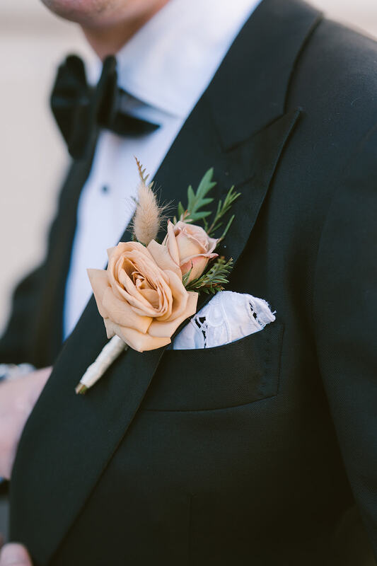 A dashing groom in a classic black suit, crisp white shirt, and white pocket square, showcasing a close-up of his boutonniere featuring a sophisticated dusty pink rose, adding a touch of refined elegance to his ensemble.