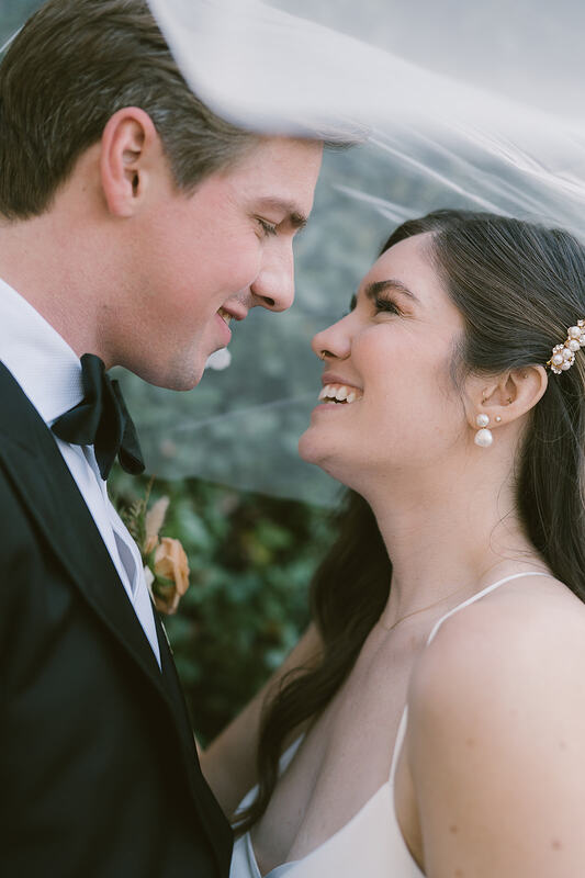A close up of a bride and groom staring lovingly at each other under the brides veil. A green hedge wall in the background. Bride has on pearl earrings, a pearl hair clip holding one side of her hair back. Groom is waring a black tux, with white undershirt, and a black bow-tie. 