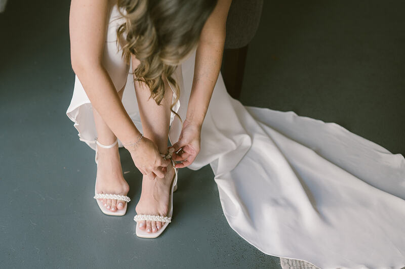 A beautiful bride, adorned in a flowing wedding dress, delicately fastening her sparkling, pearl heels in anticipation of her special day.